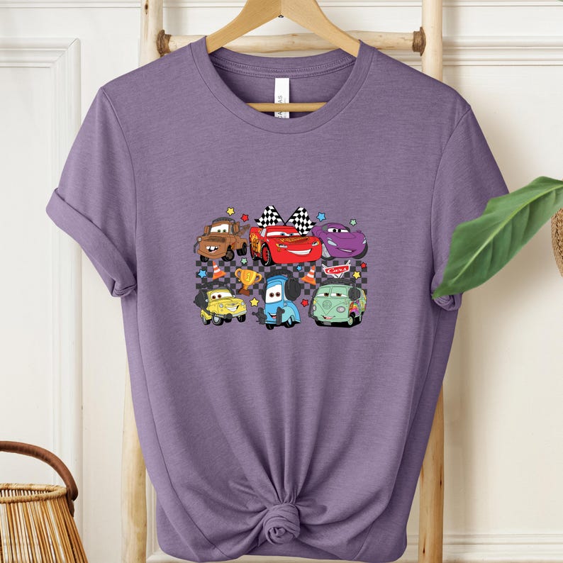 May include: A purple t-shirt featuring a colorful graphic of cartoon cars. The design includes a red race car with a checkered flag, a brown truck, and other vehicles. The cars are set against a checkered background with stars and trophies.