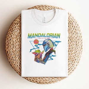 May include: White t-shirt with a colourful graphic of The Mandalorian and Grogu, with the text "MANDALORIAN" and "THIS IS THE WAY". The design includes a helmet, a sun, and a triangle. The shirt is folded on a woven surface.