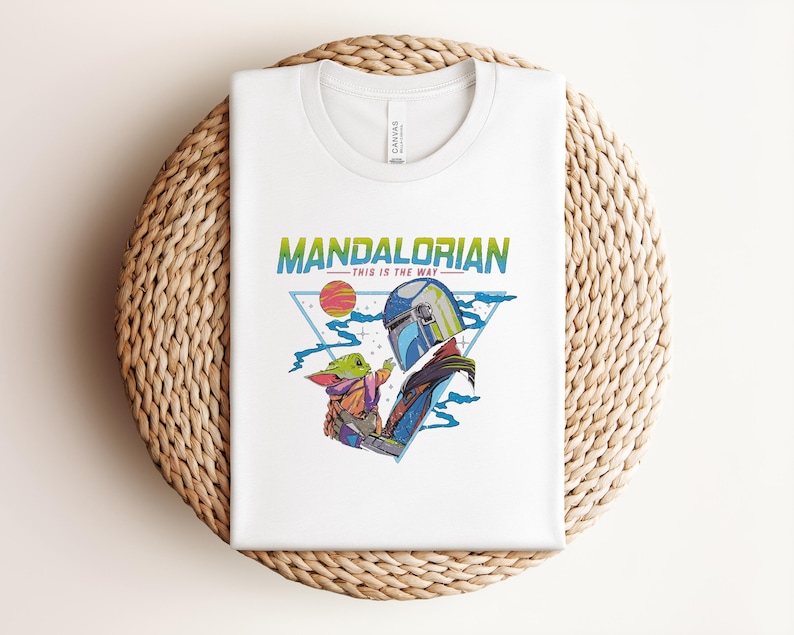 May include: White t-shirt with a colourful graphic of The Mandalorian and Grogu, with the text "MANDALORIAN" and "THIS IS THE WAY". The design includes a helmet, a sun, and a triangle. The shirt is folded on a woven surface.