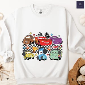 May include: White sweatshirt featuring a colorful graphic of animated cars. The design includes various car characters, checkered flags, stars, and a trophy. The Cars-themed design is centered on the front of the sweatshirt.