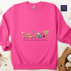 May include: A bright pink sweatshirt featuring a watercolor design of two cartoon cars, a trophy, checkered flags, and a Cozy Cone Motel sign. The text "Find your own finish line" is written below the cars.