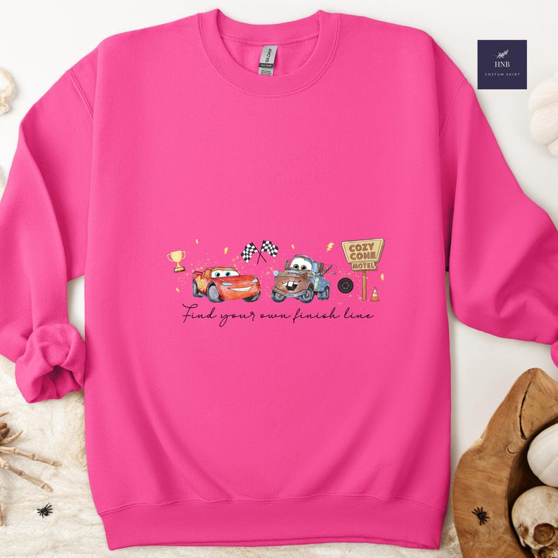 May include: A bright pink sweatshirt featuring a watercolor design of two cartoon cars, a trophy, checkered flags, and a Cozy Cone Motel sign. The text "Find your own finish line" is written below the cars.