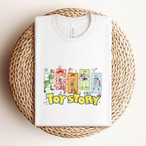 Puede incluir: Camiseta blanca con un gr&aacute;fico colorido de personajes de la pel&iacute;cula "Toy Story". El dise&ntilde;o incluye a Woody, Buzz Lightyear y otros personajes, con las palabras "Toy Story" en amarillo y azul.