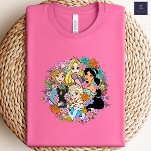 May include: A pink t-shirt featuring a colorful graphic of animated princesses surrounded by flowers. The princesses include Rapunzel, Elsa, Anna, and Jasmine. The shirt is made of a soft material and is folded on a woven surface.