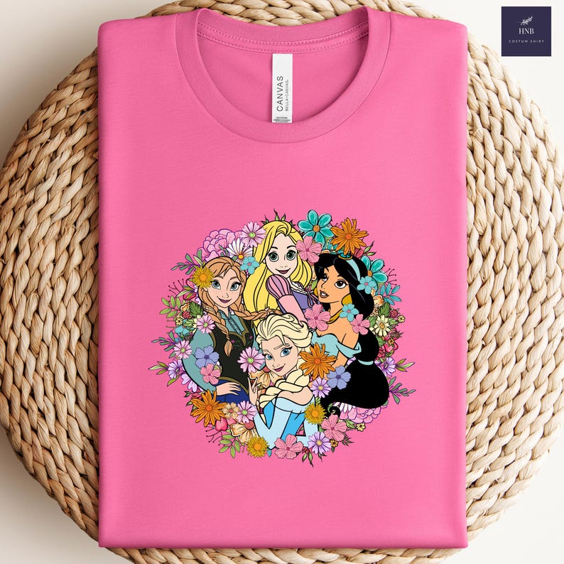 May include: A pink t-shirt featuring a colorful graphic of animated princesses surrounded by flowers. The princesses include Rapunzel, Elsa, Anna, and Jasmine. The shirt is made of a soft material and is folded on a woven surface.
