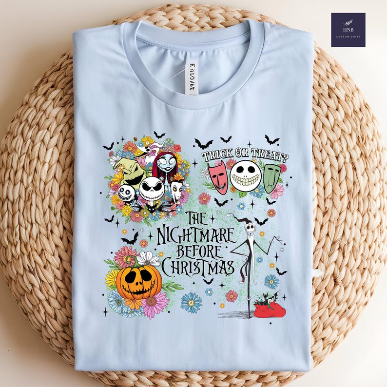 May include: Light blue t-shirt featuring a colorful graphic from "The Nightmare Before Christmas." The design includes Jack Skellington, Sally, Zero, and other characters, along with the text "Trick or Treat?" and "The Nightmare Before Christmas."