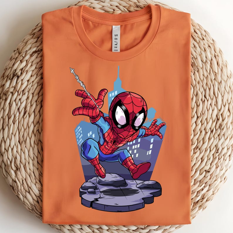 May include: Orange t-shirt featuring a cartoon Spider-Man graphic. The superhero is depicted in his classic red and blue suit, mid-action with a web, against a cityscape backdrop. The shirt is folded on a woven surface.