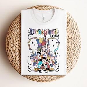 Może przedstawiać: Biały t-shirt z kolorową grafiką zamku Disneyland oraz klasycznymi postaciami z bajek, w tym Myszką Miki, Minnie, Goofym, Kaczorem Donaldem i Daisy. Tekst głosi Disneyland Est. 1955 z dodatkiem motyw&oacute;w gwiazdek.