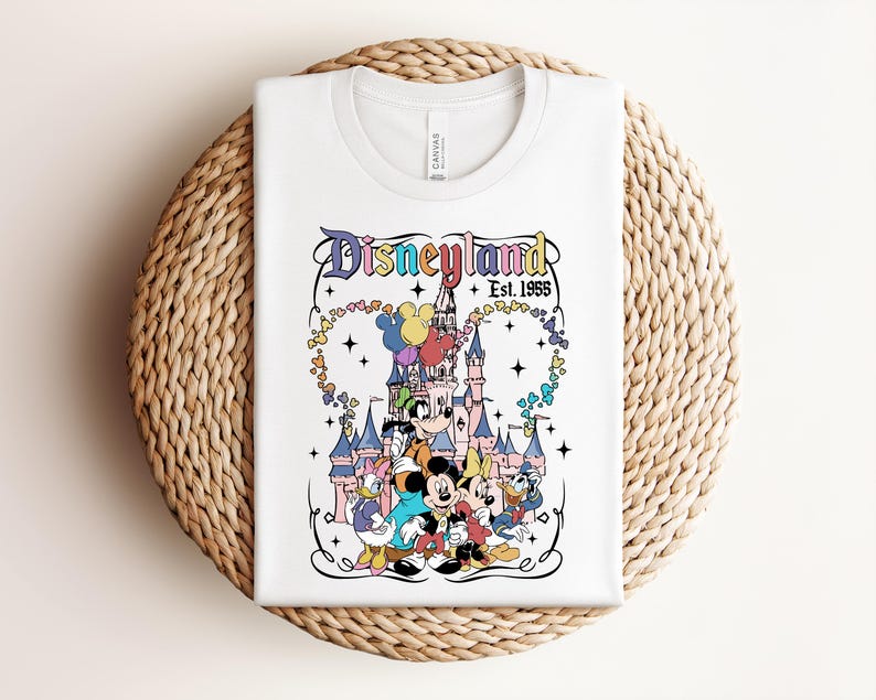 Może przedstawiać: Biały t-shirt z kolorową grafiką zamku Disneyland oraz klasycznymi postaciami z bajek, w tym Myszką Miki, Minnie, Goofym, Kaczorem Donaldem i Daisy. Tekst głosi Disneyland Est. 1955 z dodatkiem motyw&oacute;w gwiazdek.