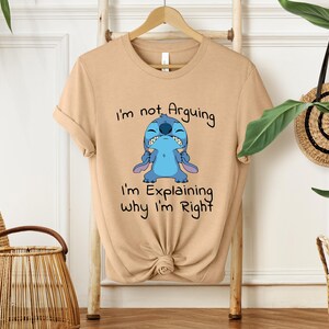 May include: A tan t-shirt featuring a blue cartoon character with the text "I'm not Arguing, I'm Explaining Why I'm Right." The character has a grumpy expression. The shirt is knotted at the bottom.