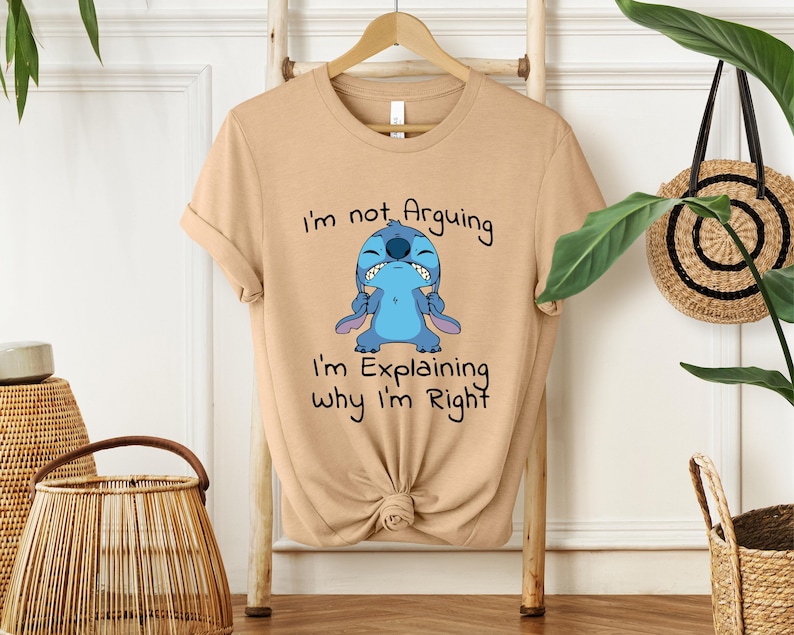 May include: A tan t-shirt featuring a blue cartoon character with the text "I'm not Arguing, I'm Explaining Why I'm Right." The character has a grumpy expression. The shirt is knotted at the bottom.