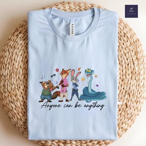 K&ouml;nnte beinhalten: Hellblaues T-Shirt mit einer Cartoon-Illustration von Zootopia-Charakteren. Das Design umfasst einen Biber, einen Fuchs, ein Kaninchen und eine Schlange, mit dem Text "Anyone can be anything" in einer Schriftart. Das T-Shirt ist auf einer gewebten Oberfl&auml;che gefaltet.