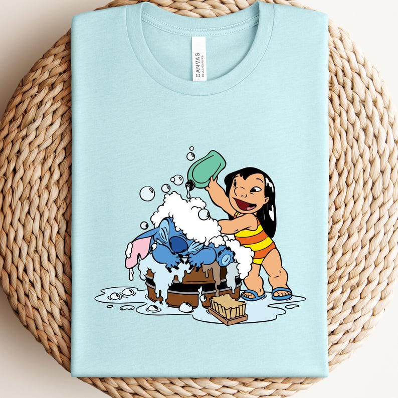 Pode incluir: Camiseta azul claro com uma ilustra&ccedil;&atilde;o de desenho animado de Lilo e Stitch. Lilo est&aacute; derramando &aacute;gua sobre Stitch, que est&aacute; em um barril de madeira cheio de bolhas. A camiseta tem gola redonda e uma etiqueta que diz "Canvas".