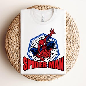 May include: White t-shirt featuring a graphic of Spider-Man in a web design, with the word "SPIDER-MAN" in red block letters. The superhero is depicted in his classic red and blue suit.