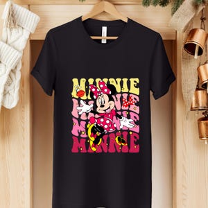 May include: Black t-shirt featuring a colorful graphic of Minnie Mouse in a polka-dot dress and bow. The design includes the name "Minnie" in a retro font with yellow, pink, and red hues. The shirt is hanging on a wooden hanger.