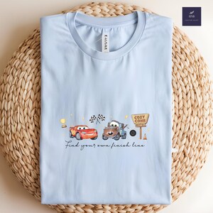 May include: Light blue t-shirt featuring a Cars-themed design. The graphic includes Lightning McQueen, Mater, a Cozy Cone Motel sign, and the text "Find your own finish line." The shirt is folded on a woven surface.