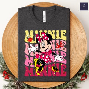 May include: Dark gray t-shirt featuring a colorful graphic of Minnie Mouse with the word "MINNIE" stacked in retro font. Minnie is wearing a red polka dot dress and bow. The shirt is displayed on a wooden plate with greenery.