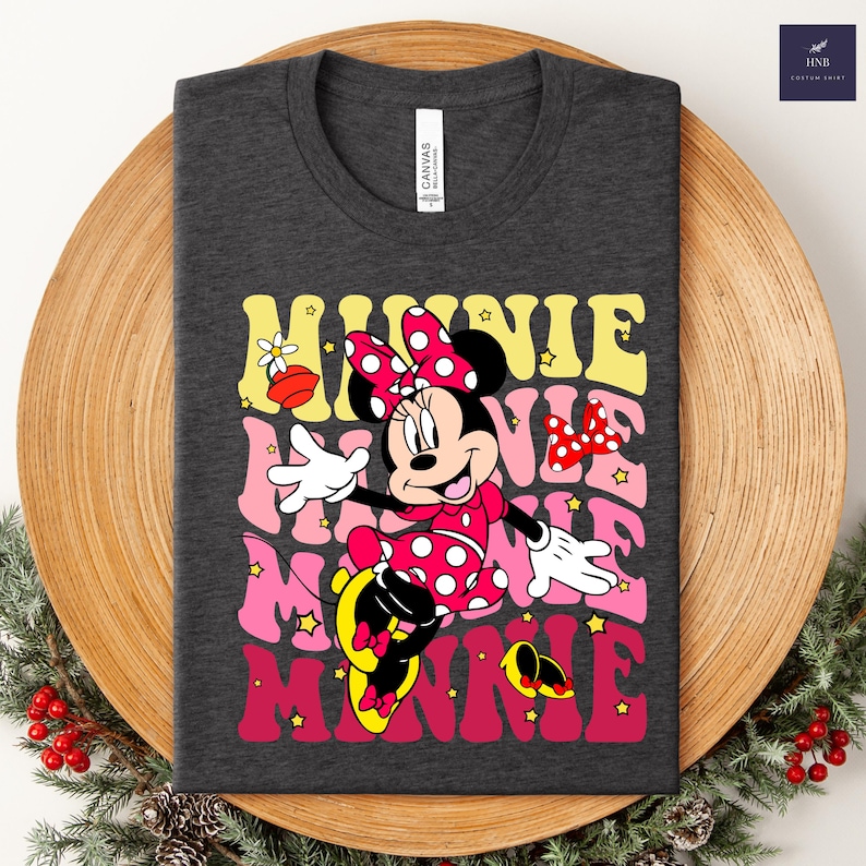 May include: Dark gray t-shirt featuring a colorful graphic of Minnie Mouse with the word "MINNIE" stacked in retro font. Minnie is wearing a red polka dot dress and bow. The shirt is displayed on a wooden plate with greenery.