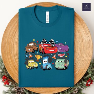 May include: Teal-colored t-shirt featuring a colorful graphic of cartoon cars. The design includes Lightning McQueen, Mater, and other characters, along with checkered flags, stars, and a trophy. The word "CARS" is also visible.