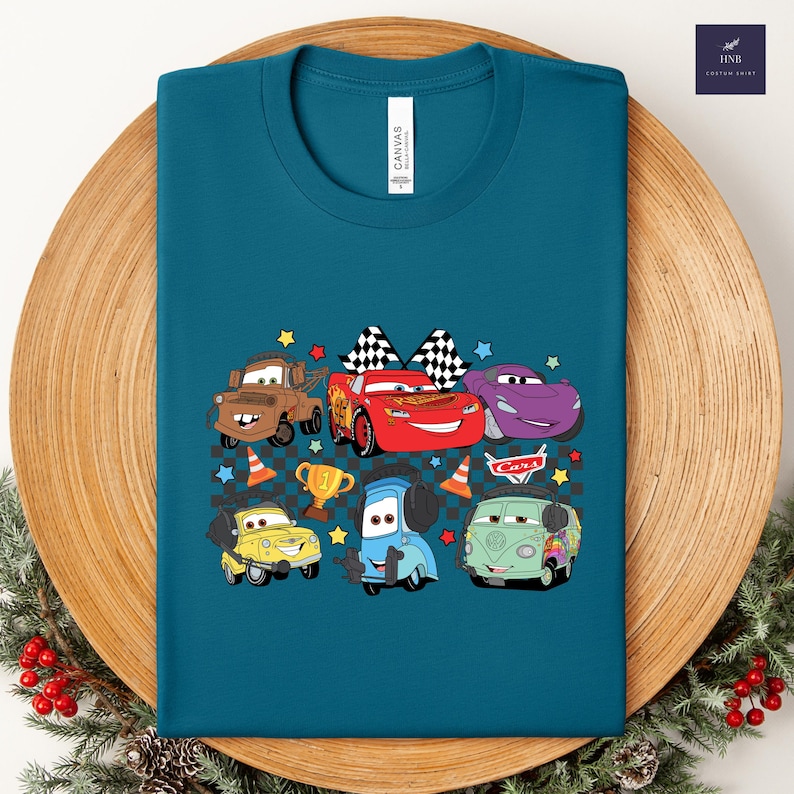 May include: Teal-colored t-shirt featuring a colorful graphic of cartoon cars. The design includes Lightning McQueen, Mater, and other characters, along with checkered flags, stars, and a trophy. The word "CARS" is also visible.