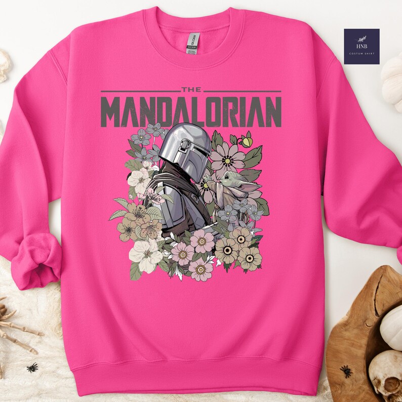 Puede incluir: Una sudadera rosa intenso con un dise&ntilde;o de The Mandalorian y Grogu rodeados de flores. El texto "THE MANDALORIAN" est&aacute; impreso en gris. La sudadera tiene mangas largas y cuello redondo.