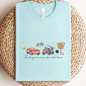 May include: Light blue t-shirt featuring a watercolor design with two cartoon cars, a trophy, checkered flags, and a motel sign. The text "Find your own finish line" is written below the cars.