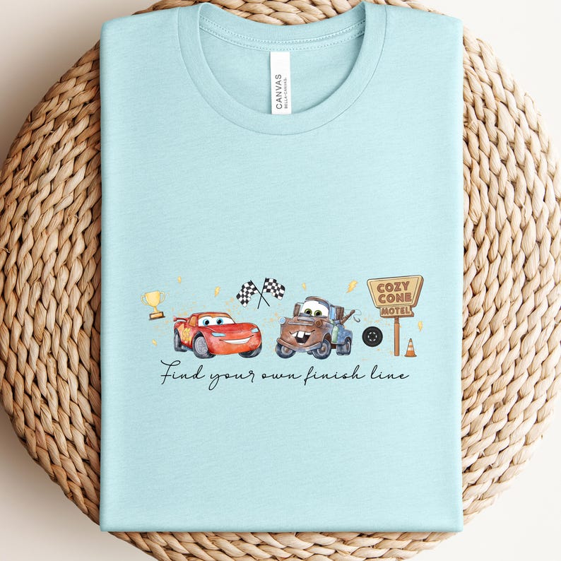 May include: Light blue t-shirt featuring a watercolor design with two cartoon cars, a trophy, checkered flags, and a motel sign. The text "Find your own finish line" is written below the cars.