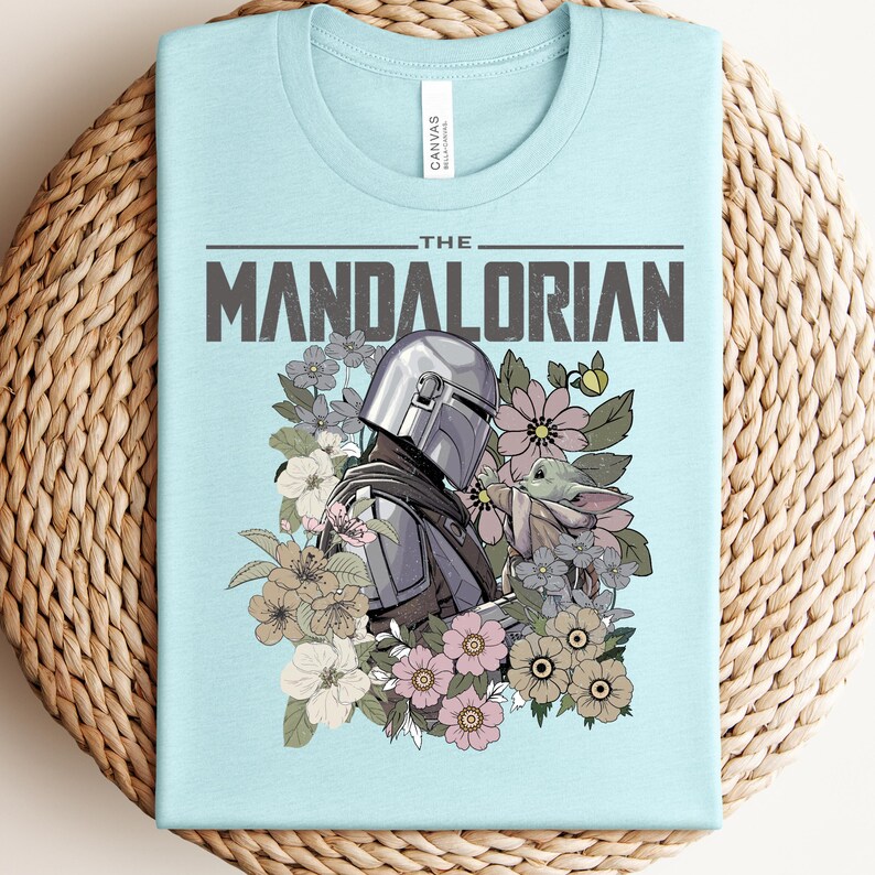 Puede incluir: Camiseta azul claro con un dise&ntilde;o de The Mandalorian y Grogu rodeados de flores. El texto "THE MANDALORIAN" est&aacute; impreso en la parte superior. La camiseta est&aacute; hecha de un material suave y tiene cuello redondo.