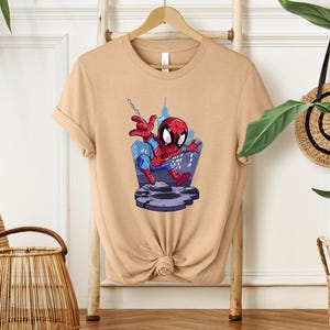 May include: A tan t-shirt featuring a cartoon illustration of Spider-Man. The superhero is depicted in his classic red and blue suit, swinging from a web in front of a cityscape. The design is centered on the shirt.