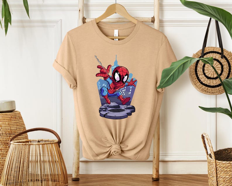 May include: A tan t-shirt featuring a cartoon illustration of Spider-Man. The superhero is depicted in his classic red and blue suit, swinging from a web in front of a cityscape. The design is centered on the shirt.