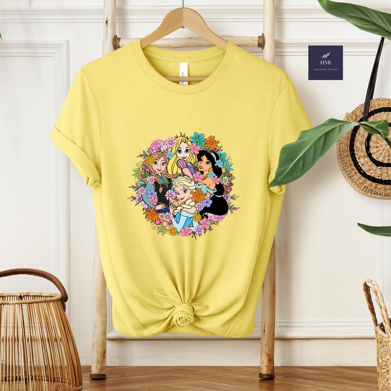 May include: Yellow t-shirt featuring a colorful graphic of animated princesses surrounded by flowers. The design includes characters with blonde, brown, and black hair. The shirt is hanging on a wooden rack.