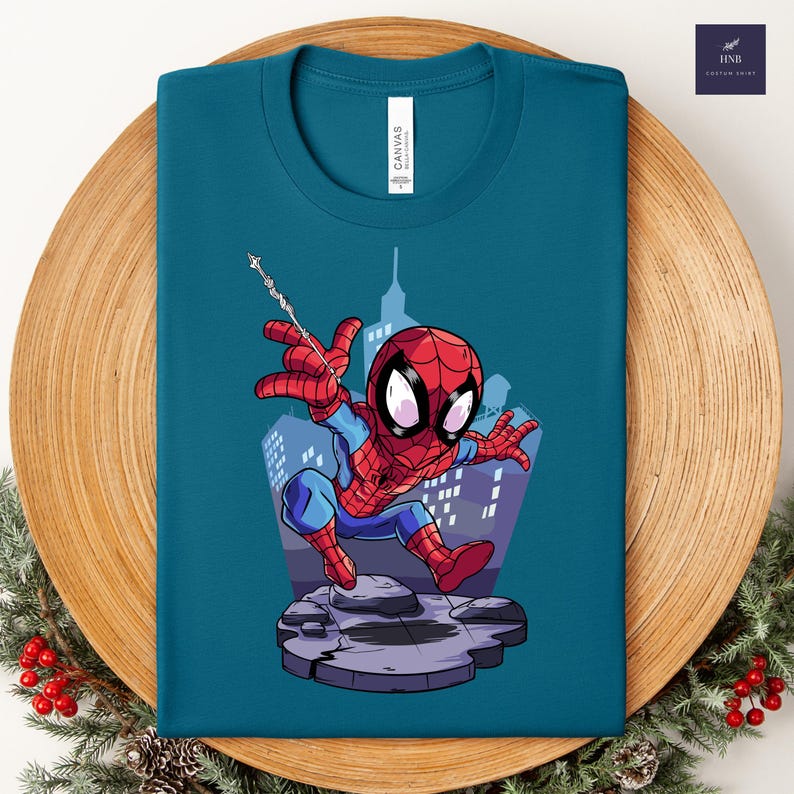 May include: Teal t-shirt featuring a cartoon Spider-Man design. The superhero is depicted in a dynamic pose with a web, set against a cityscape backdrop. The shirt has a crew neck and the brand name "CANVAS" is printed on the shirt.