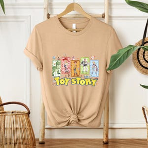 Puede incluir: Una camiseta de color beige con las palabras "Toy Story" en amarillo y un gr&aacute;fico colorido de personajes de la pel&iacute;cula animada. La camiseta se muestra en una percha de madera.