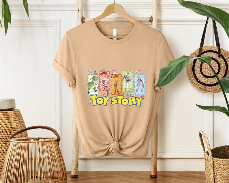 Puede incluir: Una camiseta de color beige con las palabras "Toy Story" en amarillo y un gr&aacute;fico colorido de personajes de la pel&iacute;cula animada. La camiseta se muestra en una percha de madera.