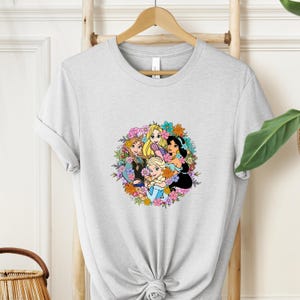 May include: A light gray t-shirt featuring a colorful floral design with cartoon princesses. The design includes Anna, Elsa, Rapunzel, and Jasmine, surrounded by a wreath of flowers. The shirt is hanging on a wooden hanger.