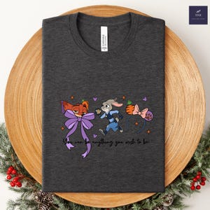 May include: Dark gray t-shirt featuring cartoon characters and the text "You can be anything you wish to be." The design includes a fox with a purple bow, a rabbit in a police uniform, and a pink bow headband. The shirt is made of canvas material.