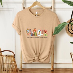 May include: A tan t-shirt featuring a whimsical illustration of cartoon animals and the text "You are my happily ever after." The design includes a fox, rabbit, and other characters, with floral accents.