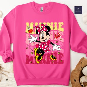 May include: A bright pink sweatshirt featuring a graphic of Minnie Mouse in a red polka-dot dress and bow. The name "MINNIE" is printed in a retro font in yellow and red. The sweatshirt has a crew neck and long sleeves.