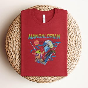 May include: A folded red t-shirt with a graphic design from the Mandalorian series. The design includes the text "MANDALORIAN" and "THIS IS THE WAY", along with illustrations of the Mandalorian and Grogu.