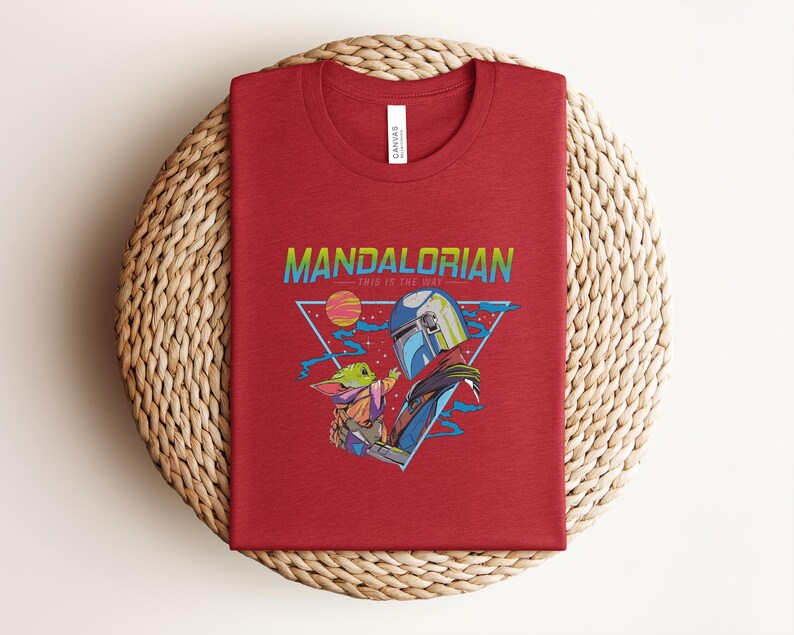 May include: A folded red t-shirt with a graphic design from the Mandalorian series. The design includes the text "MANDALORIAN" and "THIS IS THE WAY", along with illustrations of the Mandalorian and Grogu.
