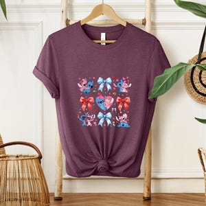 May include: A maroon t-shirt featuring a square design with cartoon characters, bows, and hearts. The design includes blue and pink characters, red and blue bows, and a heart with two characters. The shirt is hanging on a wooden hanger.