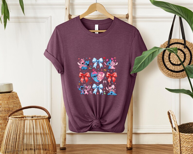 May include: A maroon t-shirt featuring a square design with cartoon characters, bows, and hearts. The design includes blue and pink characters, red and blue bows, and a heart with two characters. The shirt is hanging on a wooden hanger.