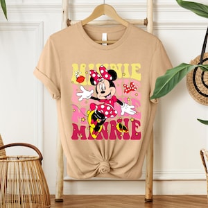 May include: Tan t-shirt featuring a colorful graphic of Minnie Mouse. Minnie is wearing a red polka dot dress and bow. The word "MINNIE" is in pink and yellow letters. The shirt is hanging on a wooden hanger.