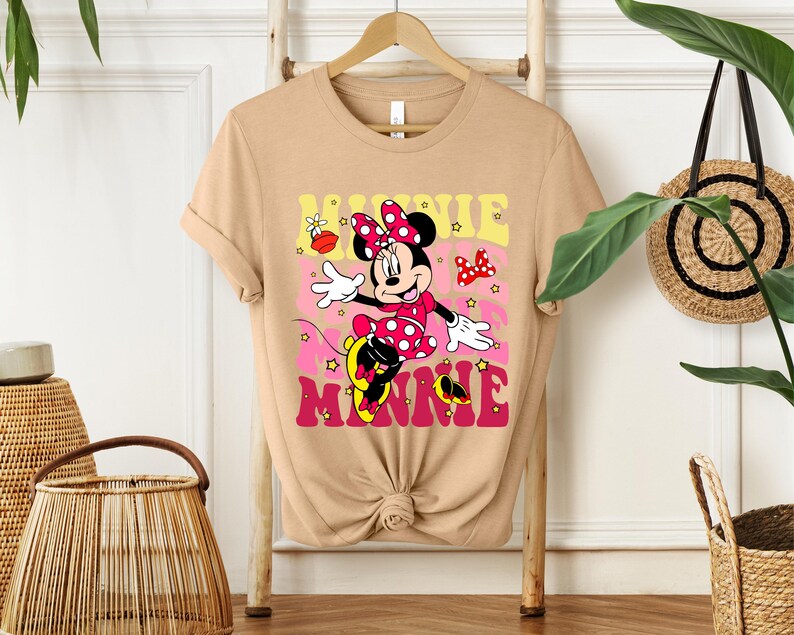May include: Tan t-shirt featuring a colorful graphic of Minnie Mouse. Minnie is wearing a red polka dot dress and bow. The word "MINNIE" is in pink and yellow letters. The shirt is hanging on a wooden hanger.