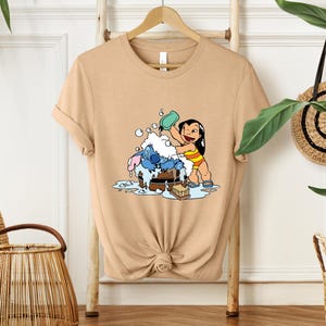Pode incluir: Camiseta bege com uma ilustra&ccedil;&atilde;o de desenho animado de Lilo e Stitch. Lilo est&aacute; despejando &aacute;gua em Stitch, que est&aacute; em uma banheira de madeira. O design inclui bolhas e uma barra de sab&atilde;o. A camiseta est&aacute; pendurada em um cabide de madeira.