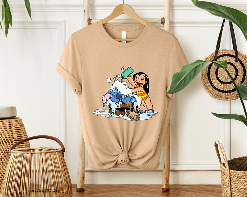Pode incluir: Camiseta bege com uma ilustra&ccedil;&atilde;o de desenho animado de Lilo e Stitch. Lilo est&aacute; despejando &aacute;gua em Stitch, que est&aacute; em uma banheira de madeira. O design inclui bolhas e uma barra de sab&atilde;o. A camiseta est&aacute; pendurada em um cabide de madeira.