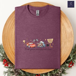 May include: A maroon t-shirt featuring a graphic of two cartoon cars racing, with a Cozy Cone Motel sign and the text "Find your own finish line." The design includes a trophy and checkered flags.