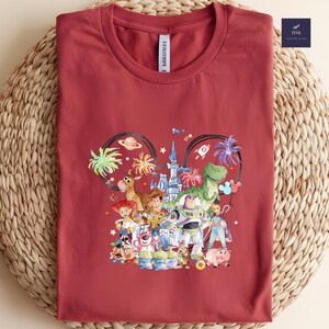 May include: A maroon t-shirt featuring a colorful graphic of various cartoon characters. The design includes a castle, a rocket, and several characters. The shirt has a round neck and a tag that says "HNB Custom Shirt".
