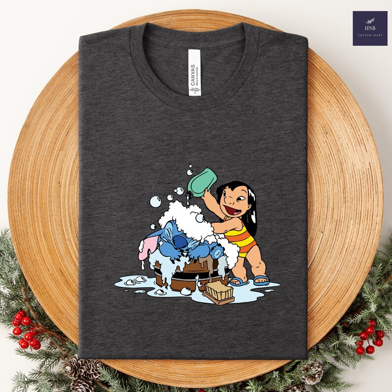 Pode incluir: Camiseta cinza escuro com uma ilustra&ccedil;&atilde;o de desenho animado de Lilo lavando Stitch em uma banheira de madeira. Lilo usa um mai&ocirc; listrado amarelo e laranja. A camisa tem o texto "Canvas" no canto superior esquerdo.
