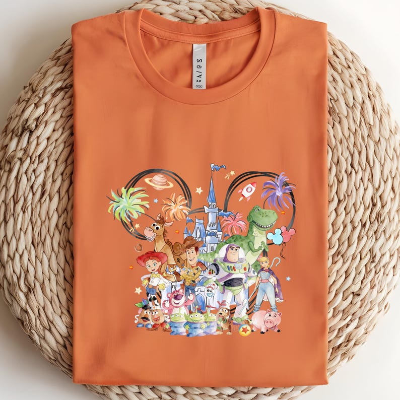 May include: Orange t-shirt featuring a colorful graphic of Disney's Toy Story characters. The design includes Woody, Buzz Lightyear, and other characters, set against a castle and palm trees. The shirt is folded on a woven surface.